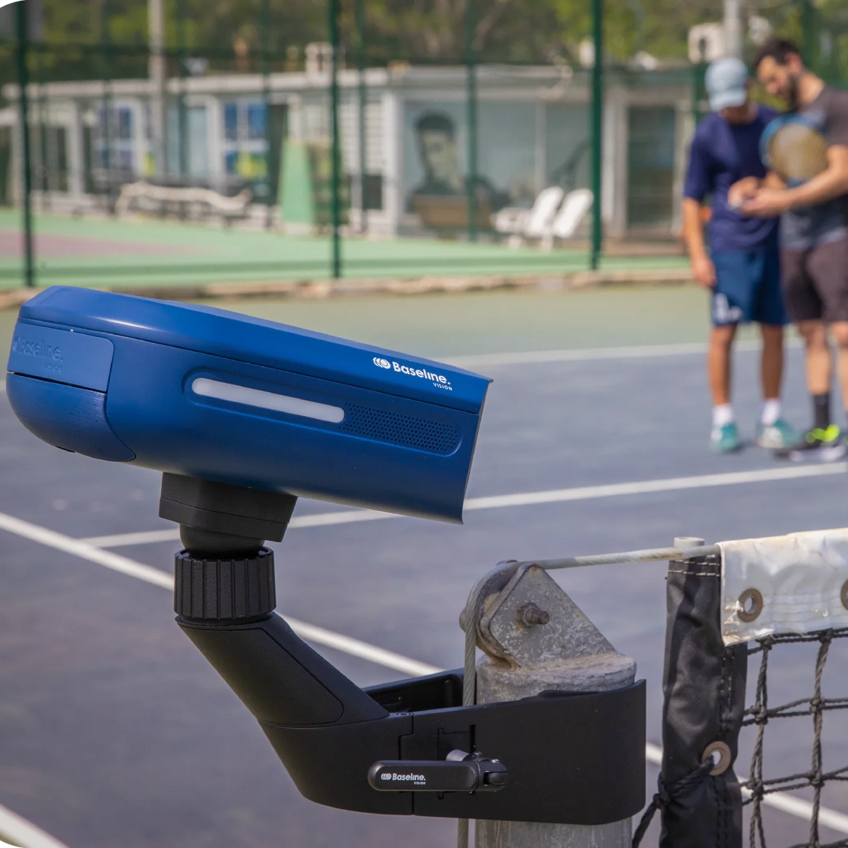 Cámara Baseline Vision con soporte en el poste de la red y, al fondo, dos jugadores en la pista