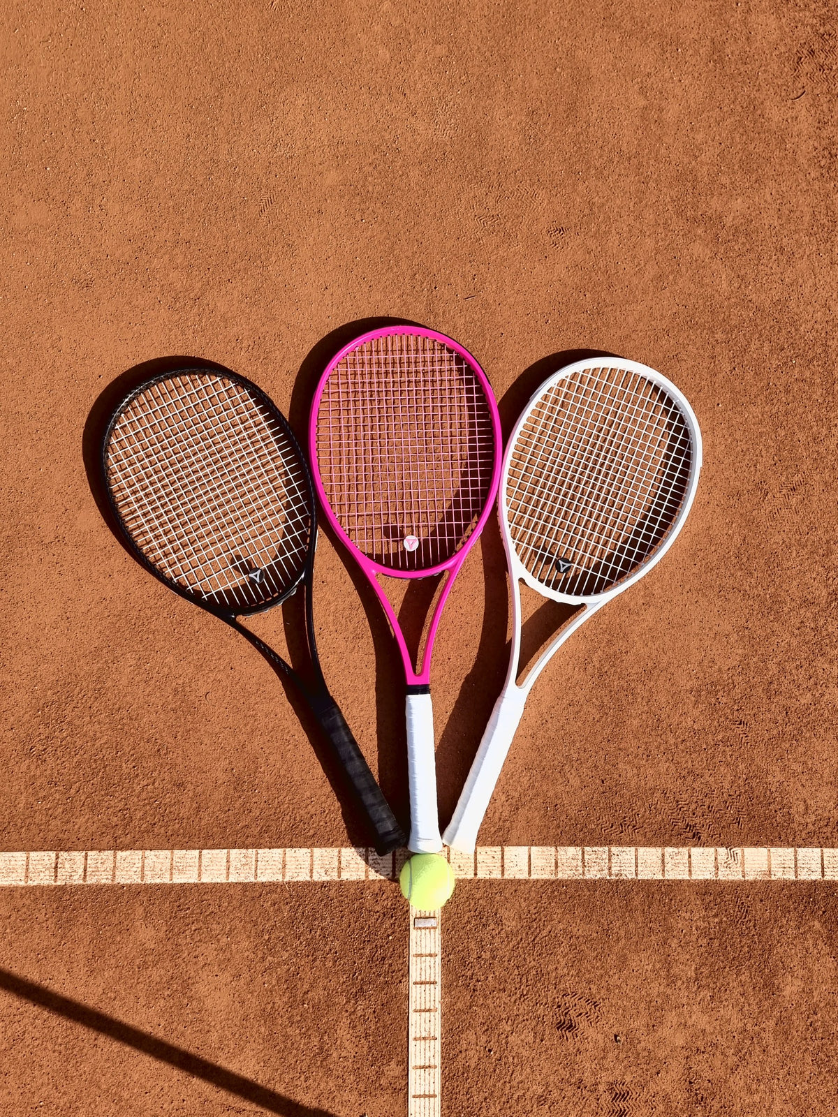 Drei myRacket-Tennisschläger in Schwarz, Pink und Weiß fächerförmig auf dem Sandplatz
