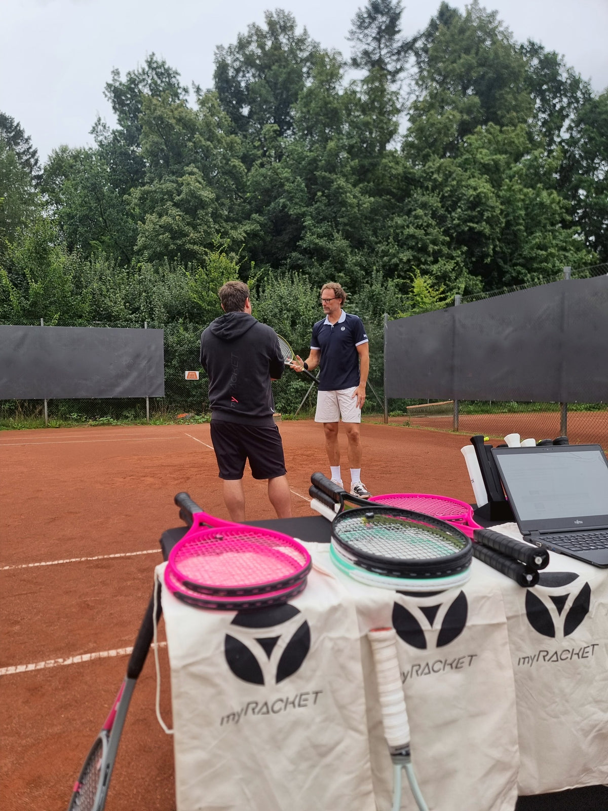 Fitter v mikine myRacket radí hráčovi na antukovom kurte, vpredu testovacie rakety na Fitting stole